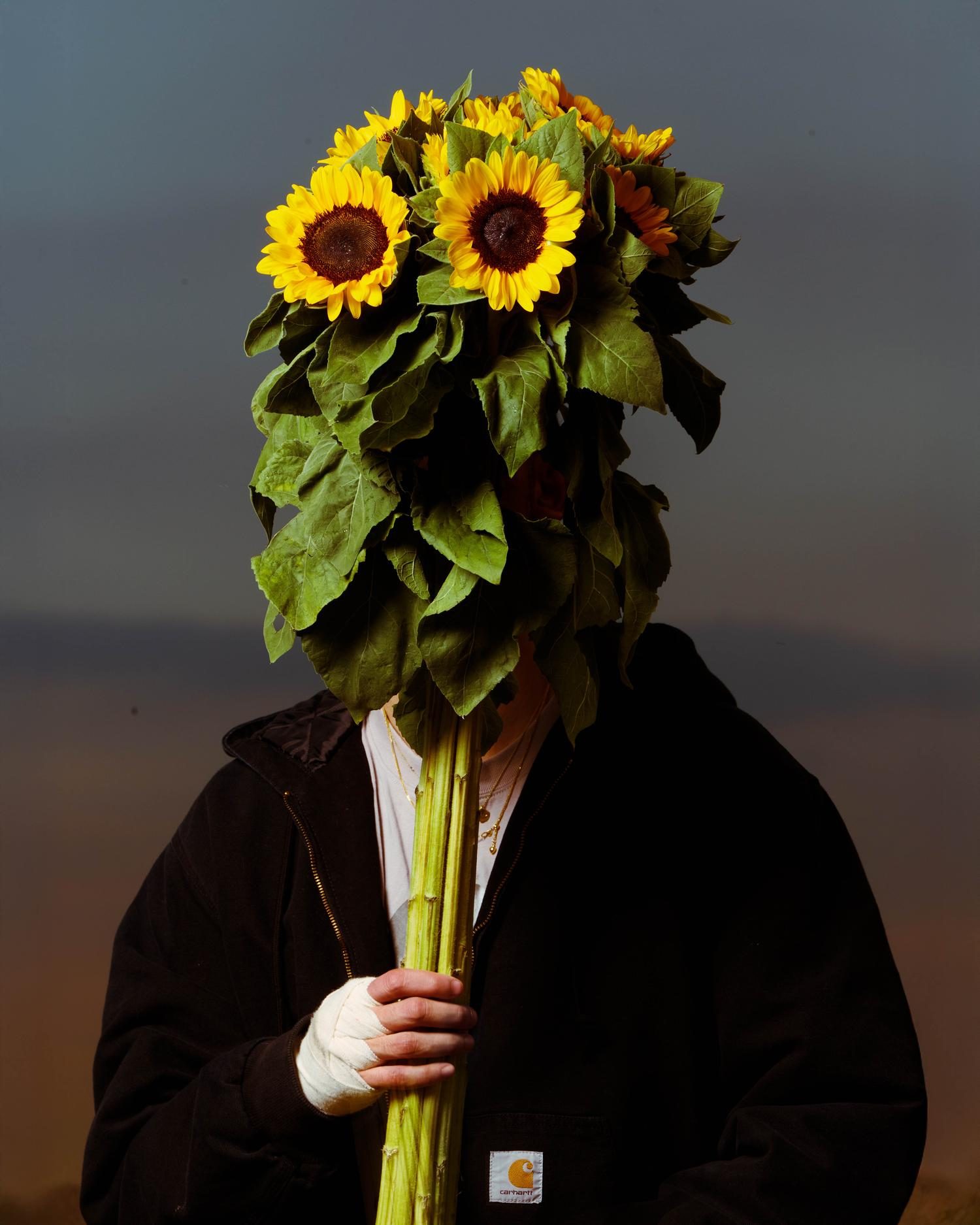Person in a black Carhartt jacket holding a bunch of sunflowers in front of their face at dusk