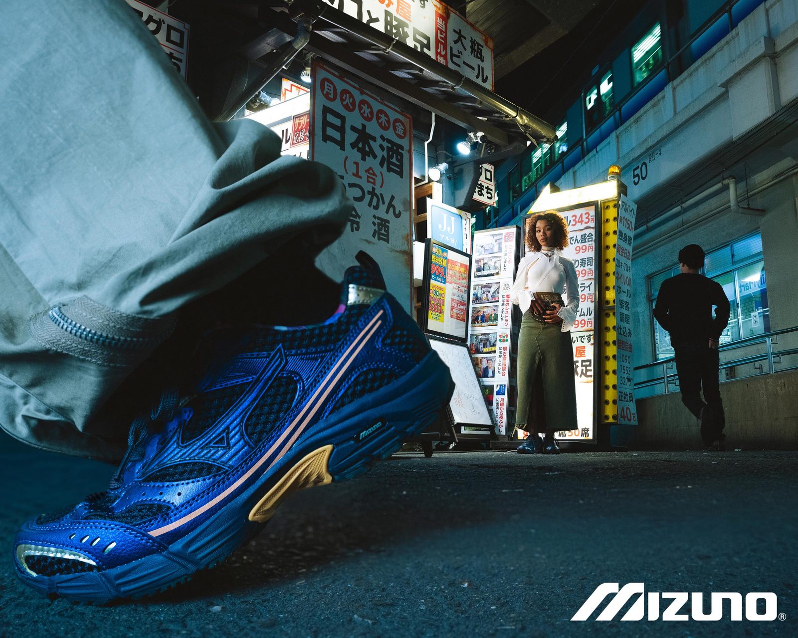 Blue Mizuno running shoe in the foreground of a neon-lit Osaka alley at night, model standing in the background