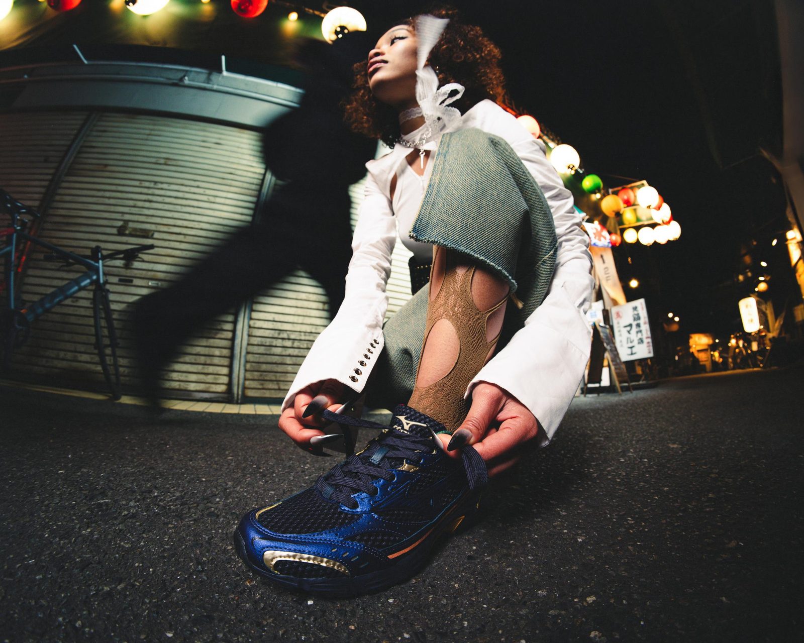 Model crouching to lace a metallic blue Mizuno sneaker under paper lanterns at night
