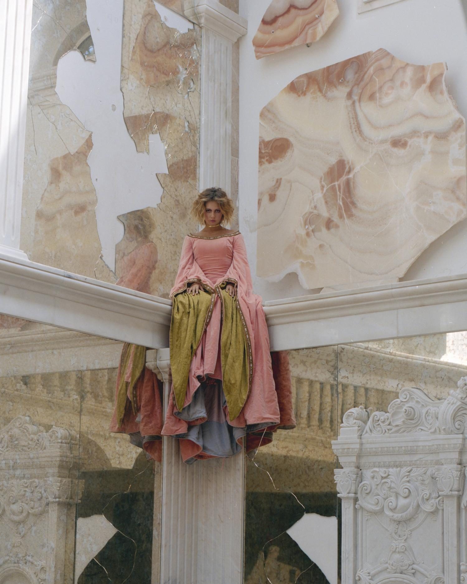 Model in a rose and gold gown seated on a marble ledge in a ruined stone interior