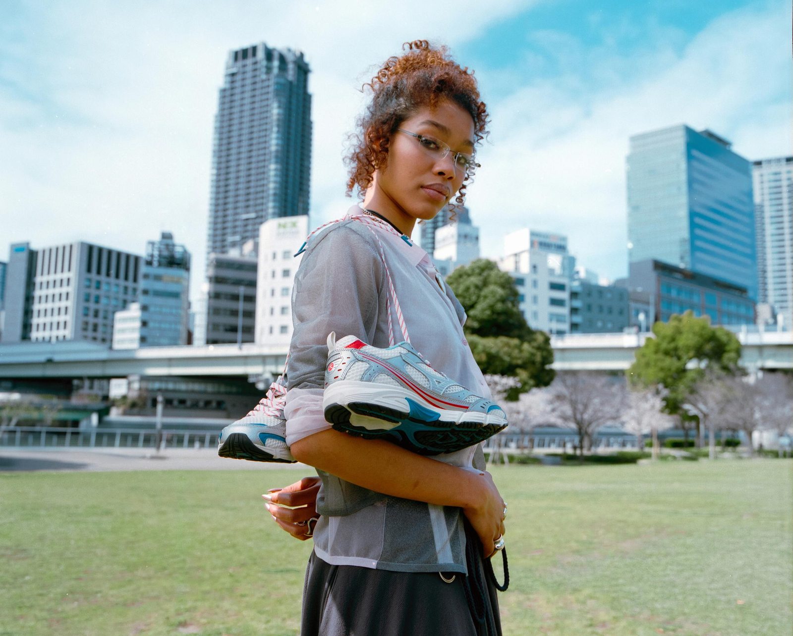 Model in a riverside park by day, carrying Mizuno sneakers under her arm, city towers behind