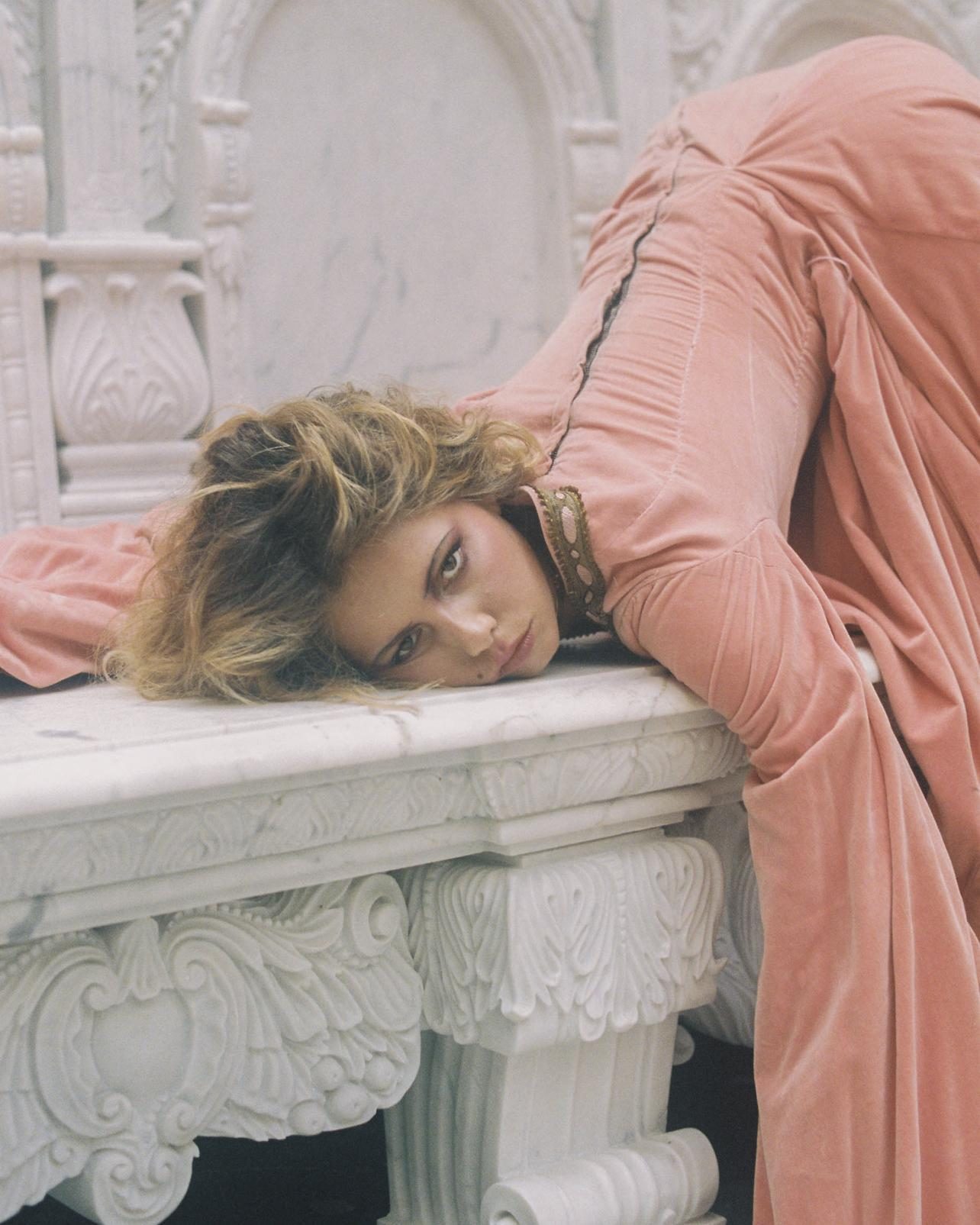 Model in a rose velvet gown resting her head on a carved marble table