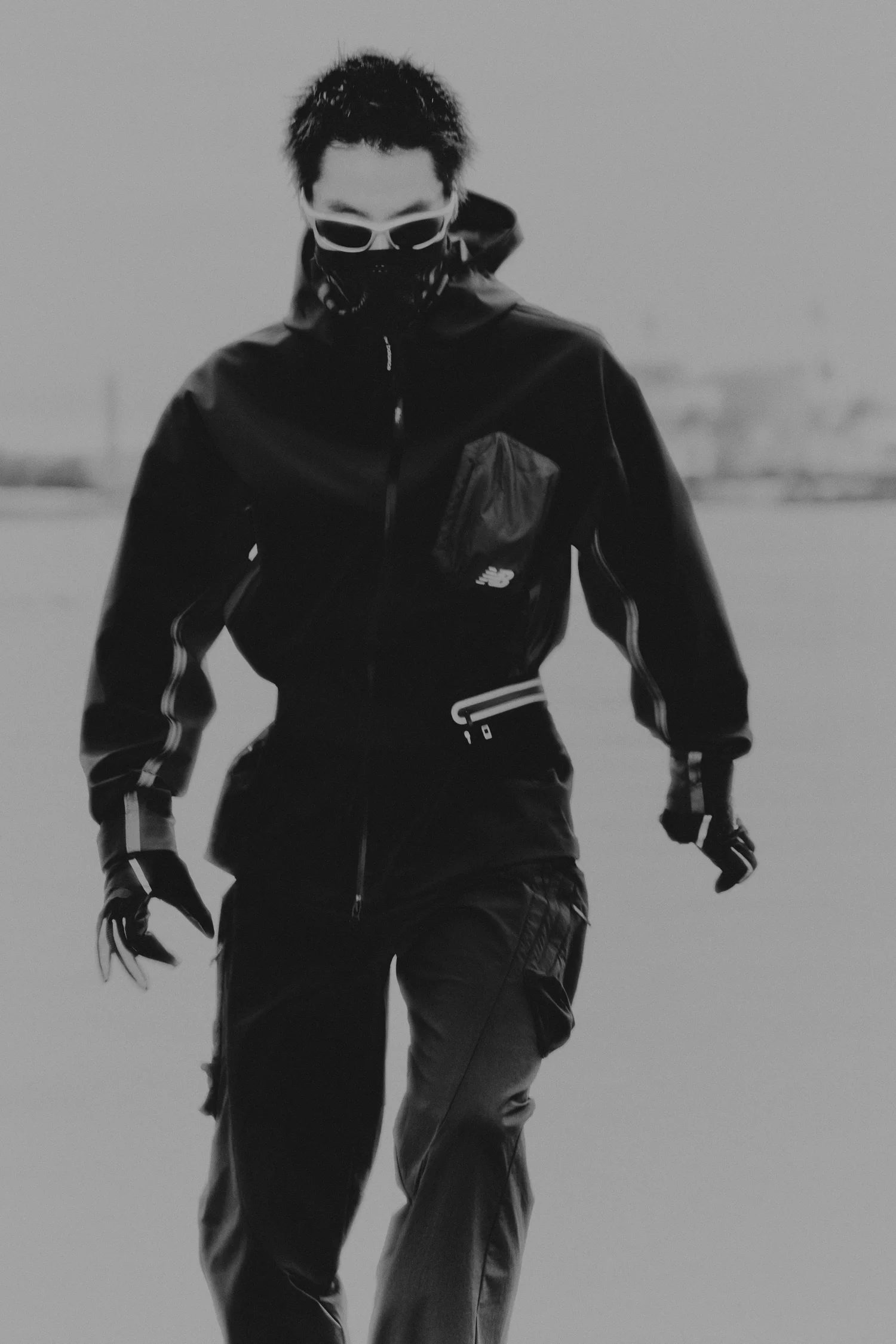 Black and white photograph of a masked runner in a New Balance shell jacket walking toward the camera