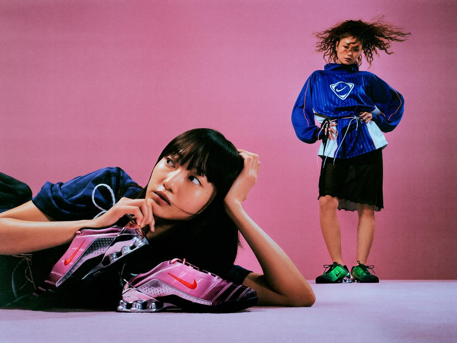 Two models in blue Nike football shirts against a pink studio backdrop, one reclining with metallic pink Shox sneakers