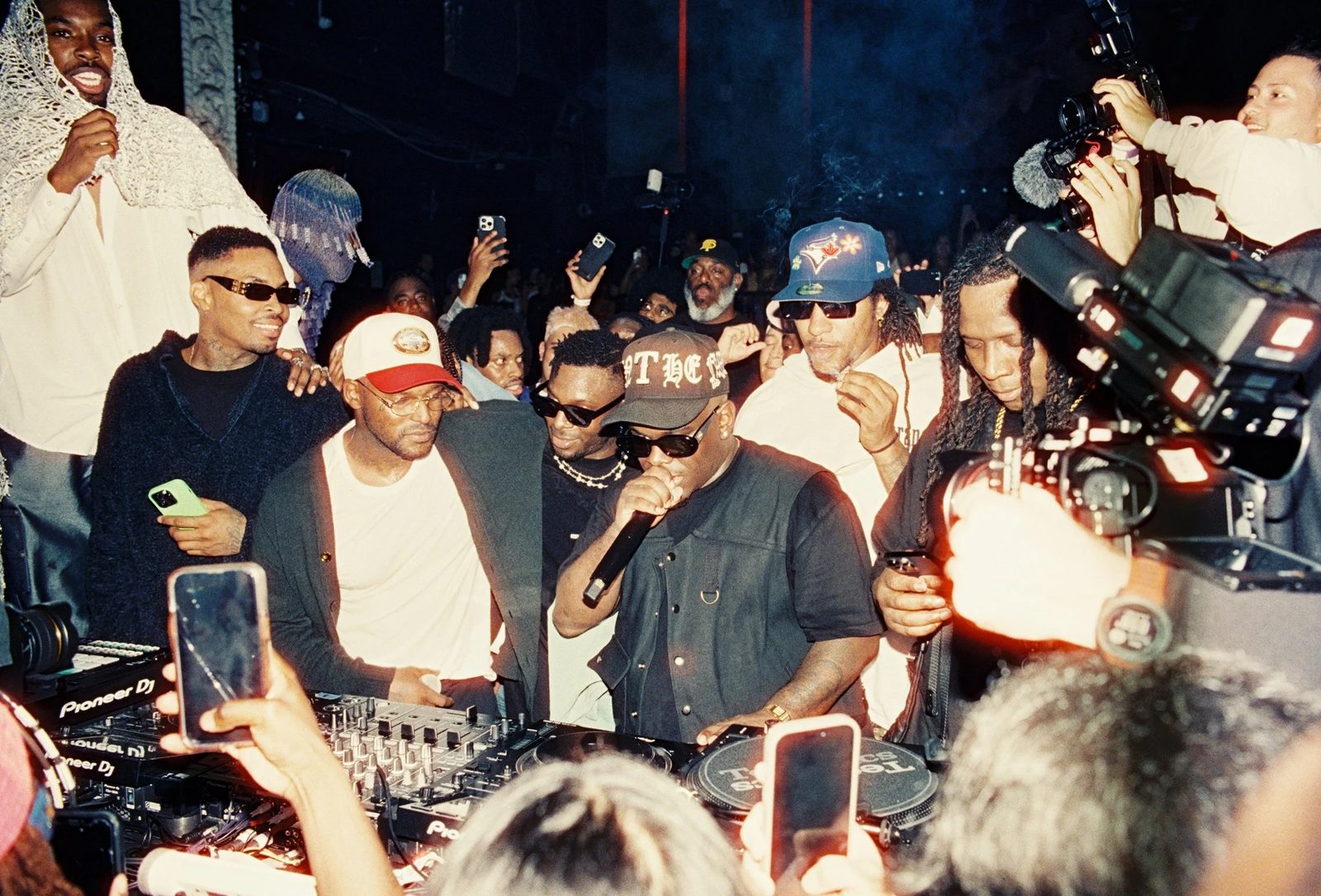 Crowded DJ booth mid-set, shot on flash film, phones and cameras raised