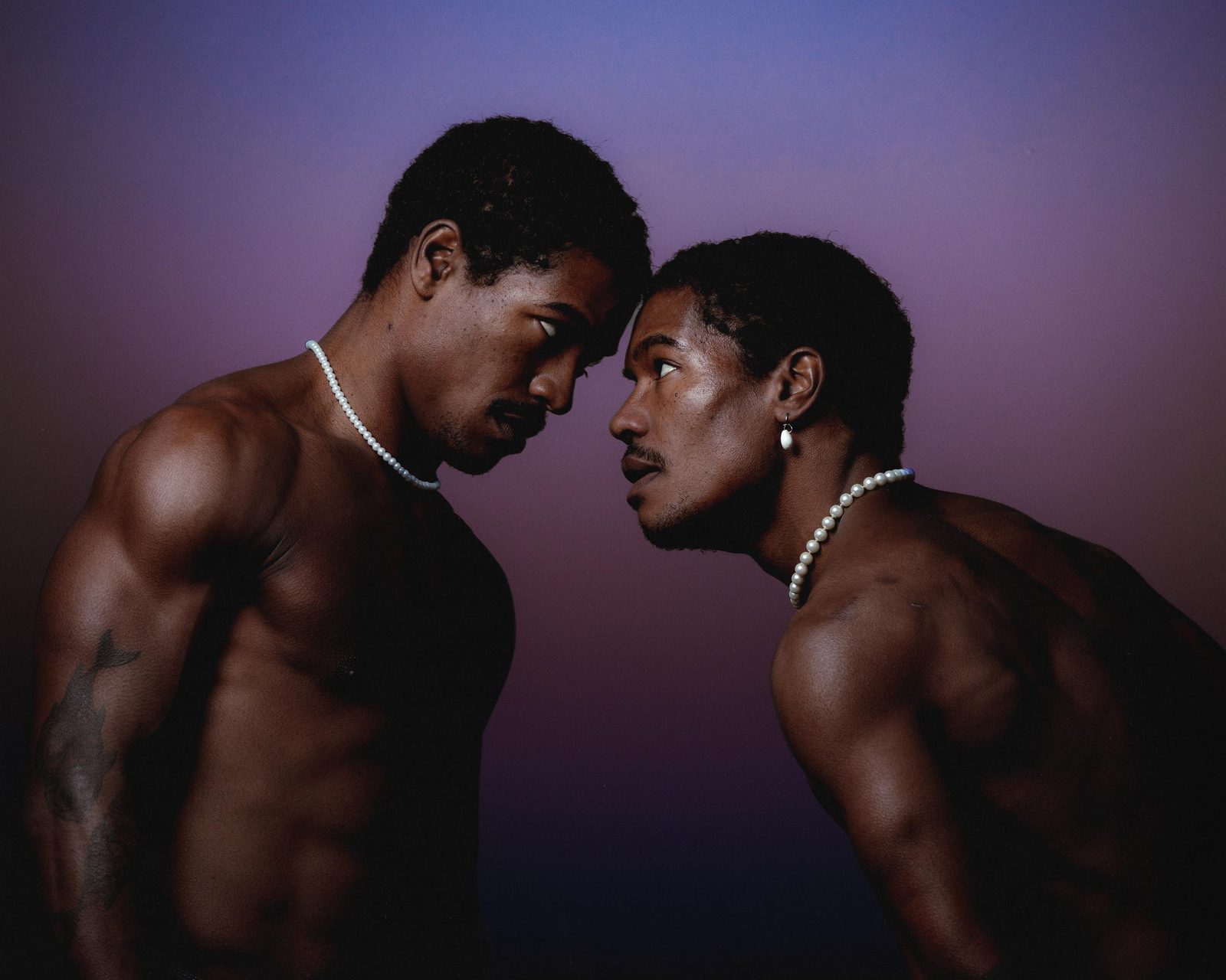 Two men wearing pearl necklaces face each other forehead to forehead against a dusk-coloured backdrop