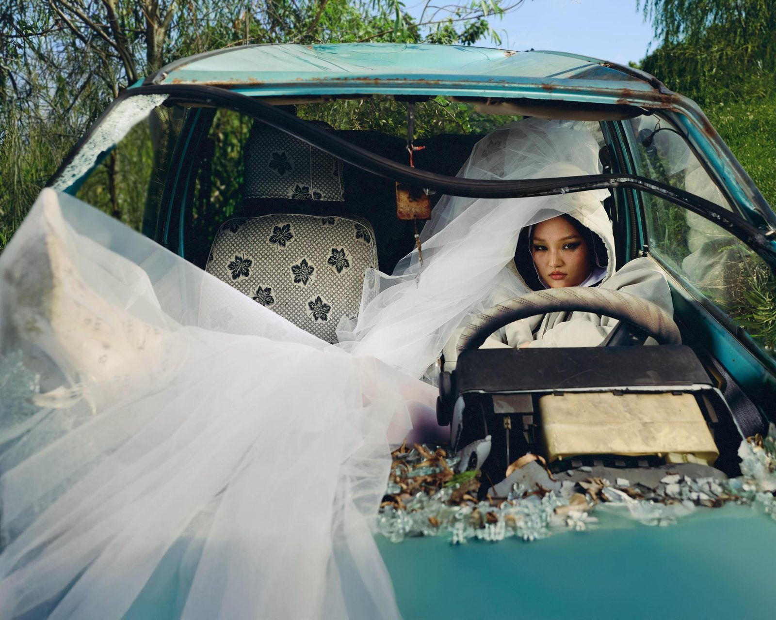Veiled model in white at the wheel of a wrecked teal car overgrown with greenery, shattered glass on the hood