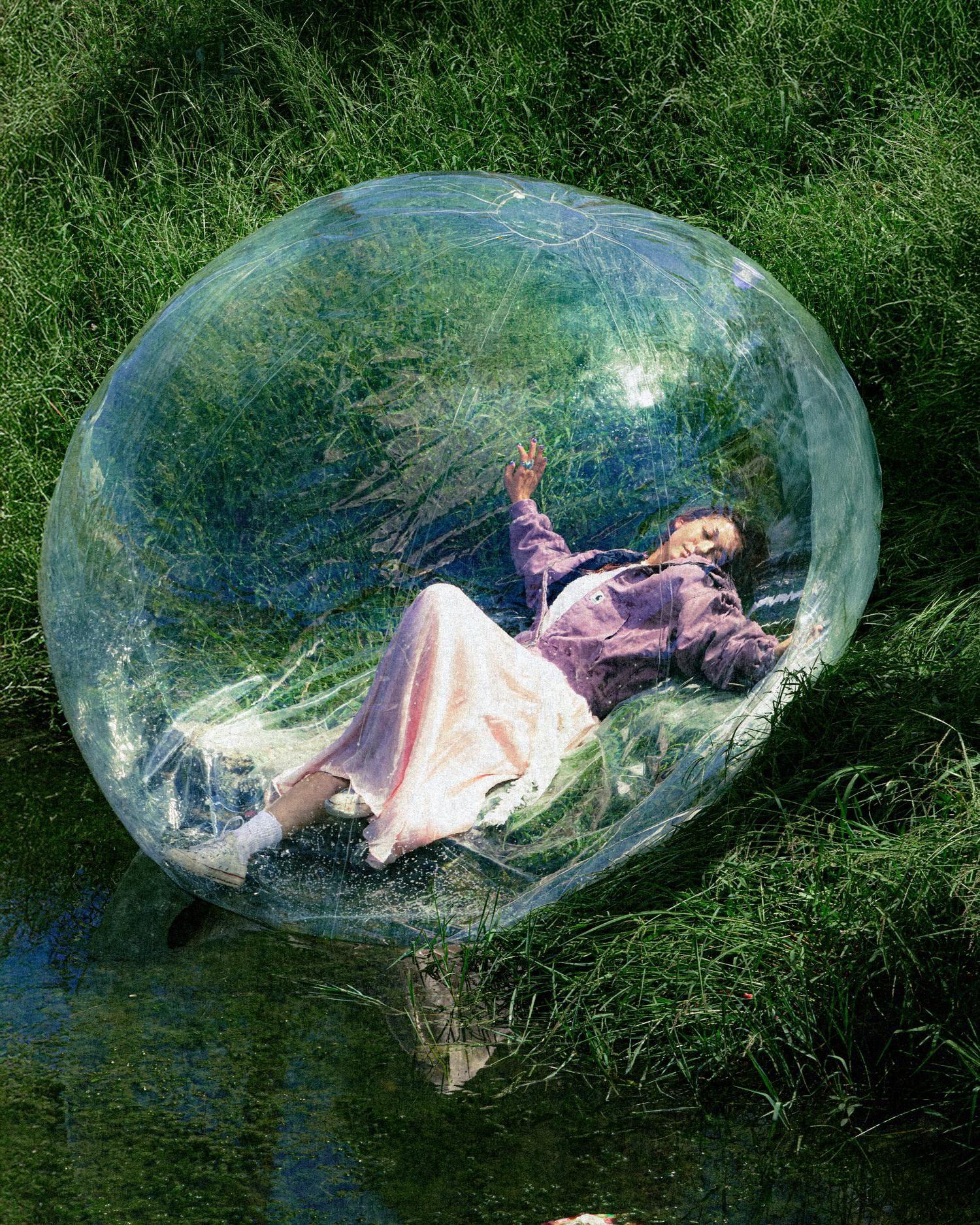 Model in a lilac Carhartt jacket lying inside a transparent inflatable sphere on a grassy riverbank